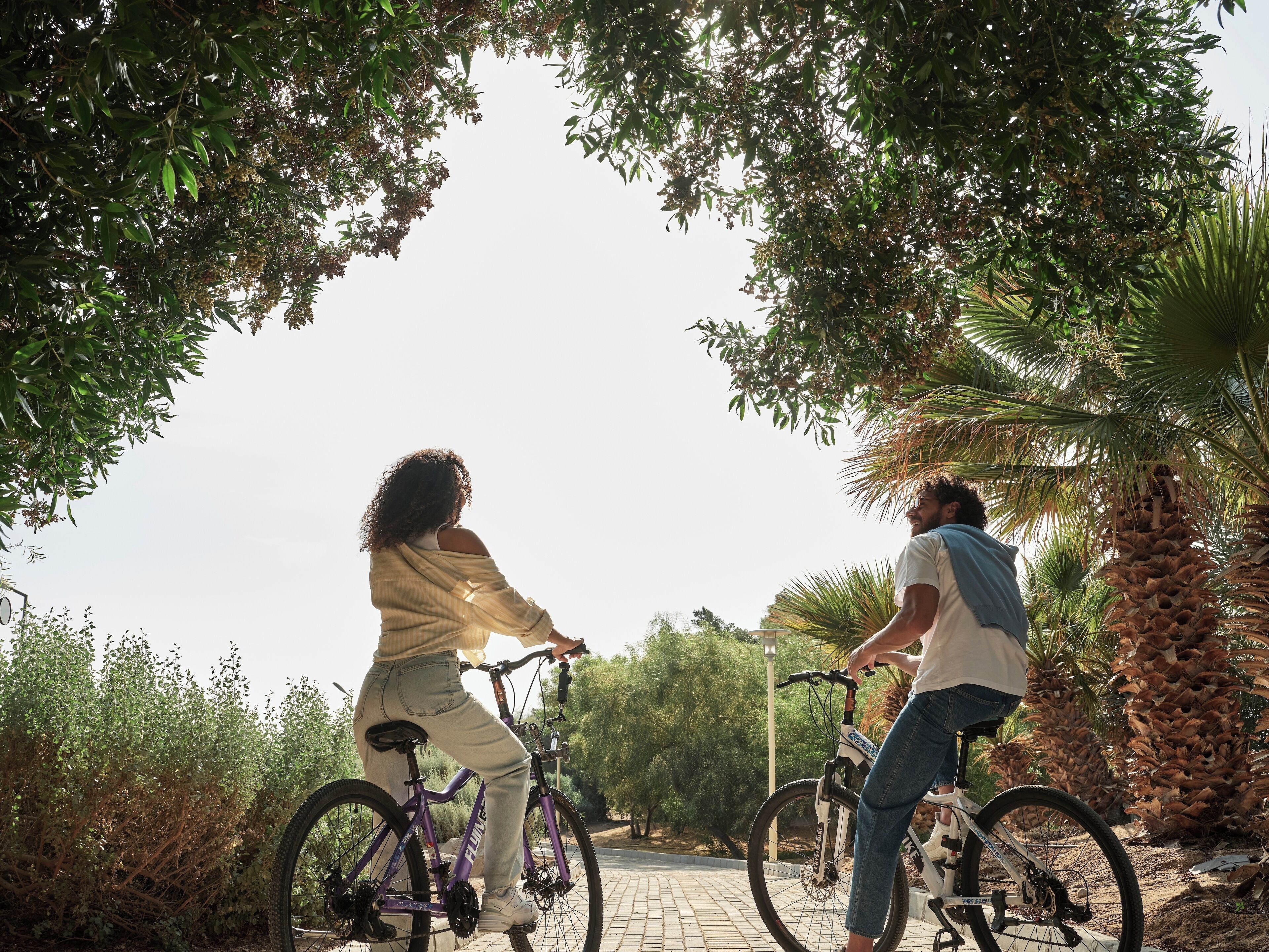 Two people riding bicycles on a paved path surrounded by trees and palm trees.