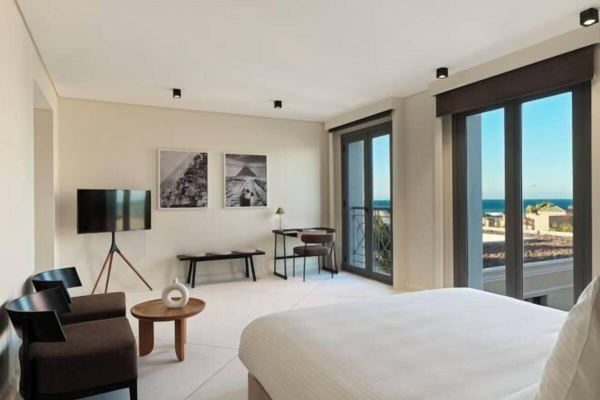 A hotel room with a bed, a desk, a chair, a TV, and a balcony with a view of the sea.