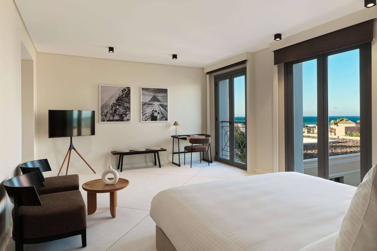 A modern hotel room with a large bed, a desk, a chair, a TV, and a balcony with a view of the sea.