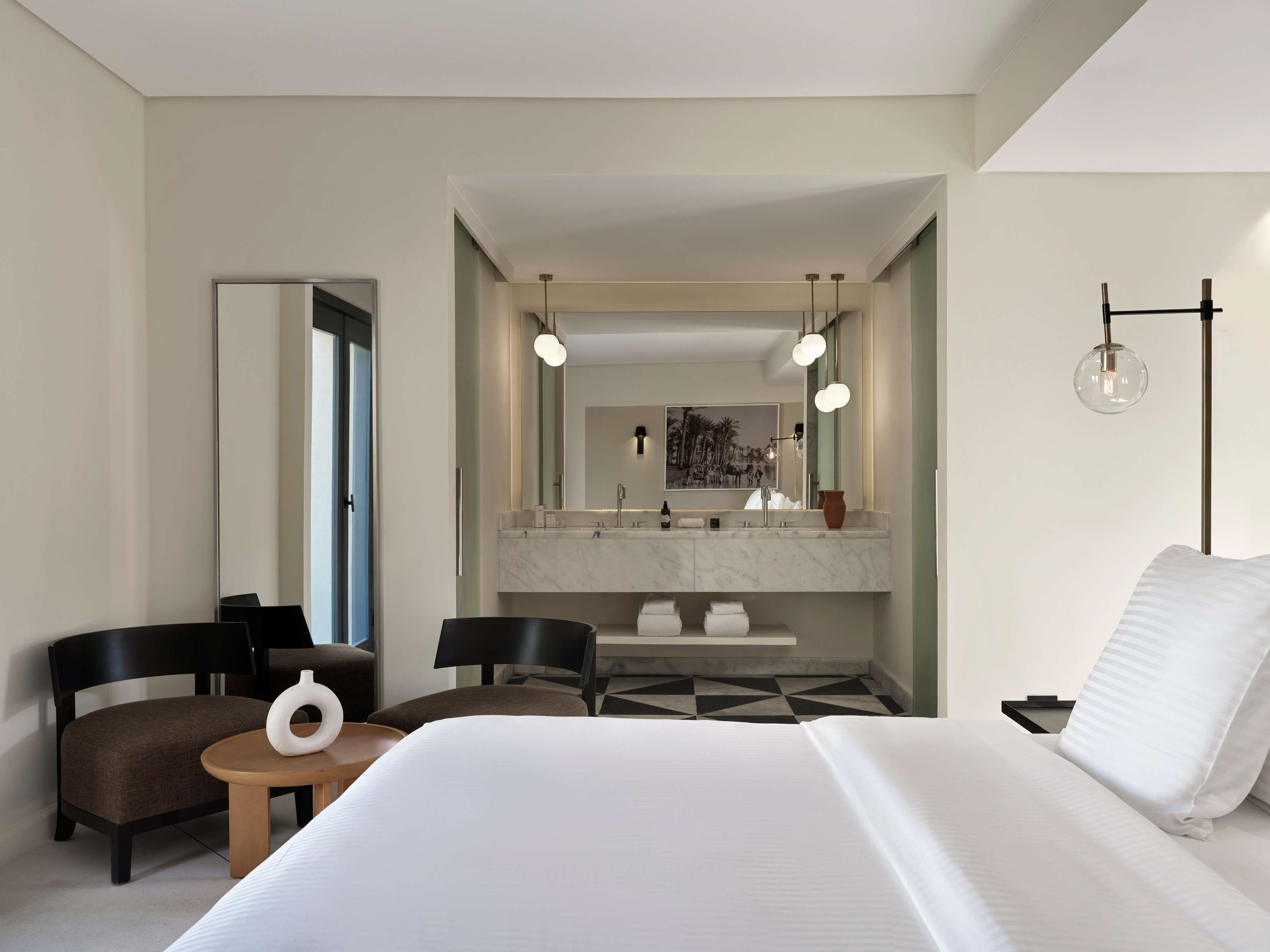 A hotel room with a large bed, a desk with two chairs, a modern bathroom with a large mirror, and a hanging light fixture.