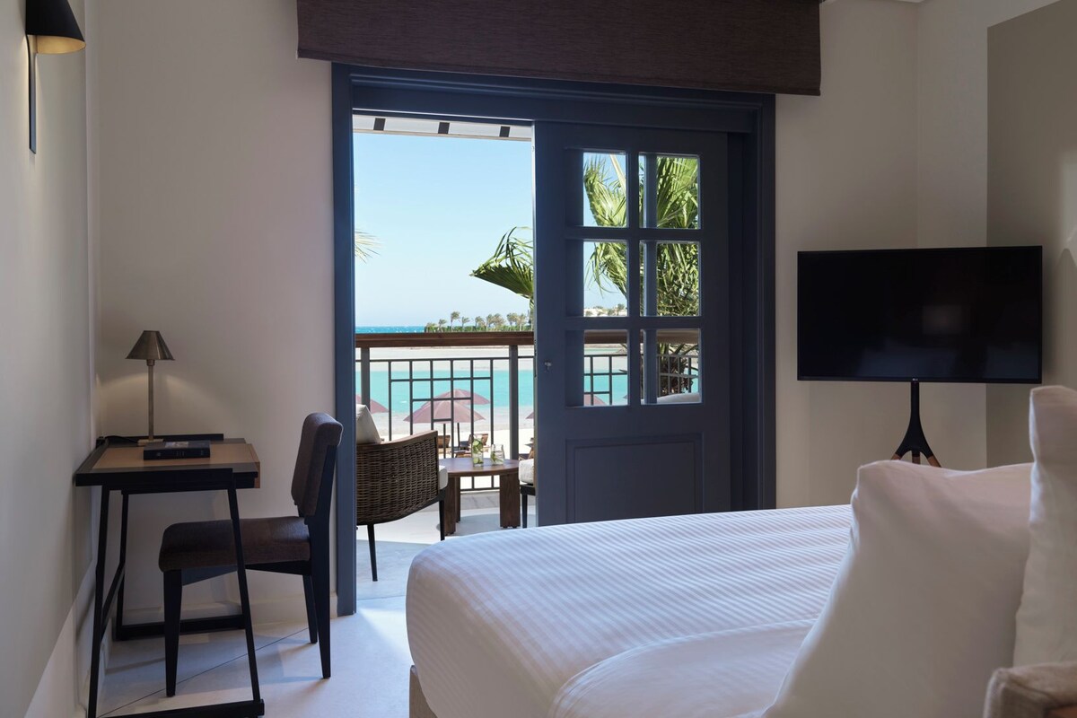 A hotel room with a bed, a desk with a lamp, a television, and a view of the beach through a door.