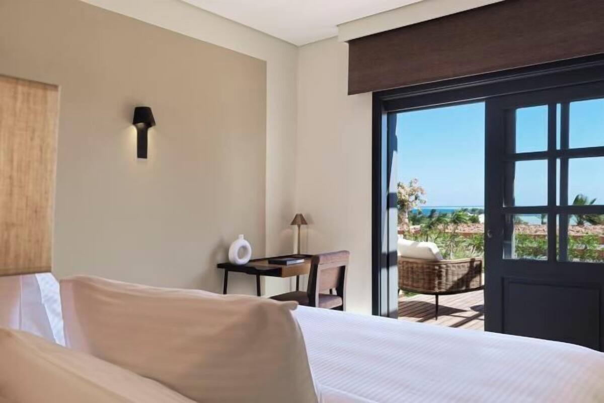 A bedroom with a bed, a desk, a chair, and a view of the ocean.