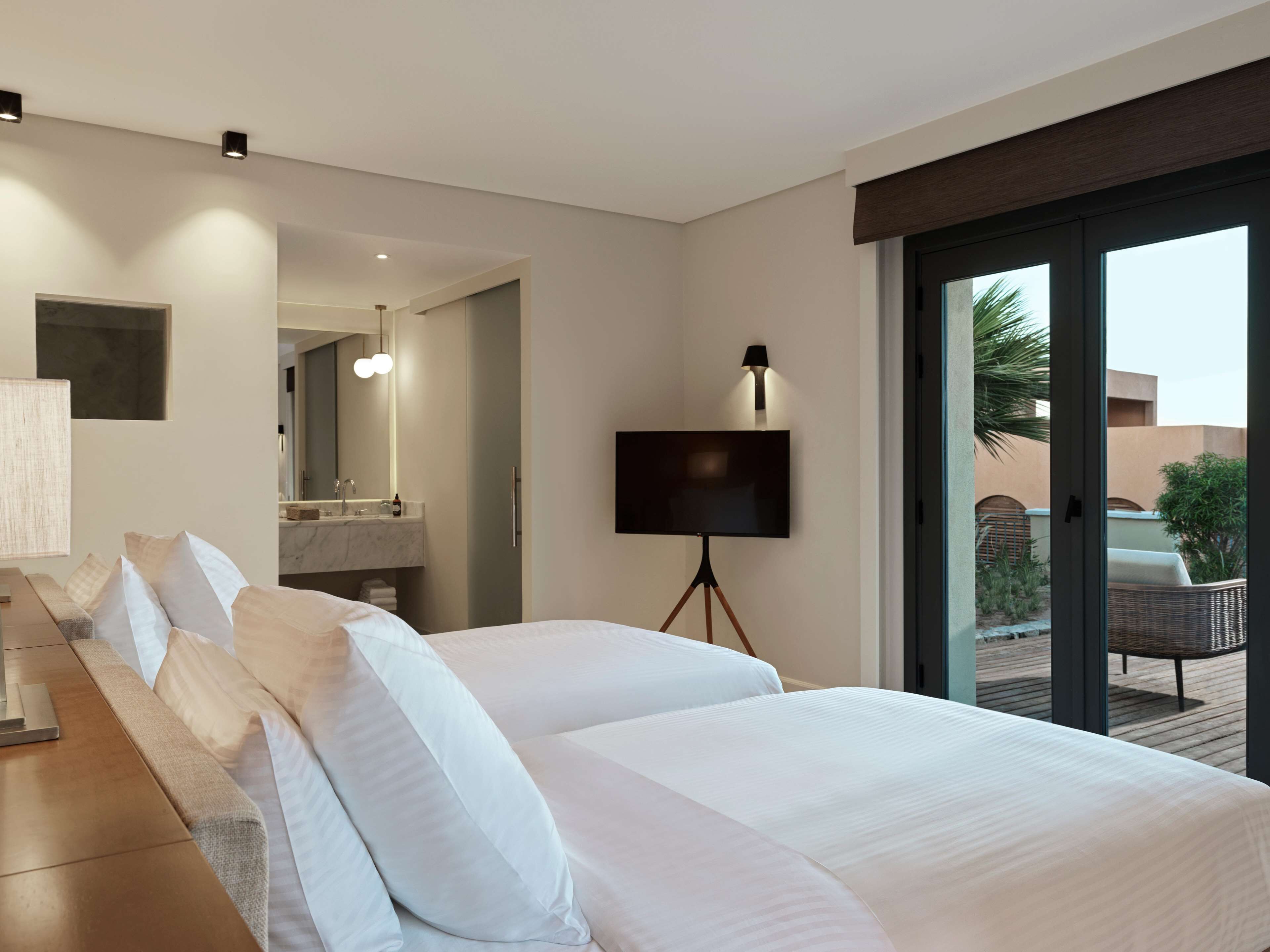 Ein modernes Hotelzimmer mit zwei Betten, einem großen Spiegel, einem Fernseher und einem Balkon mit Sitzmöbeln.