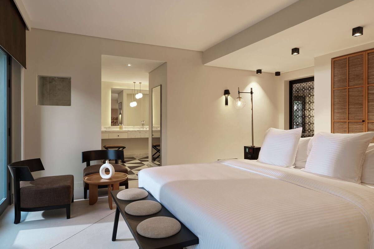 A modern hotel room with a large bed, a desk with a chair, a small round table, and a bench.