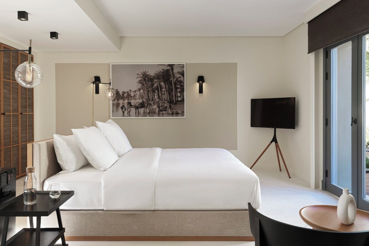A modern bedroom with a large bed, a flat-screen TV, a desk with a lamp, and a wall-mounted picture.