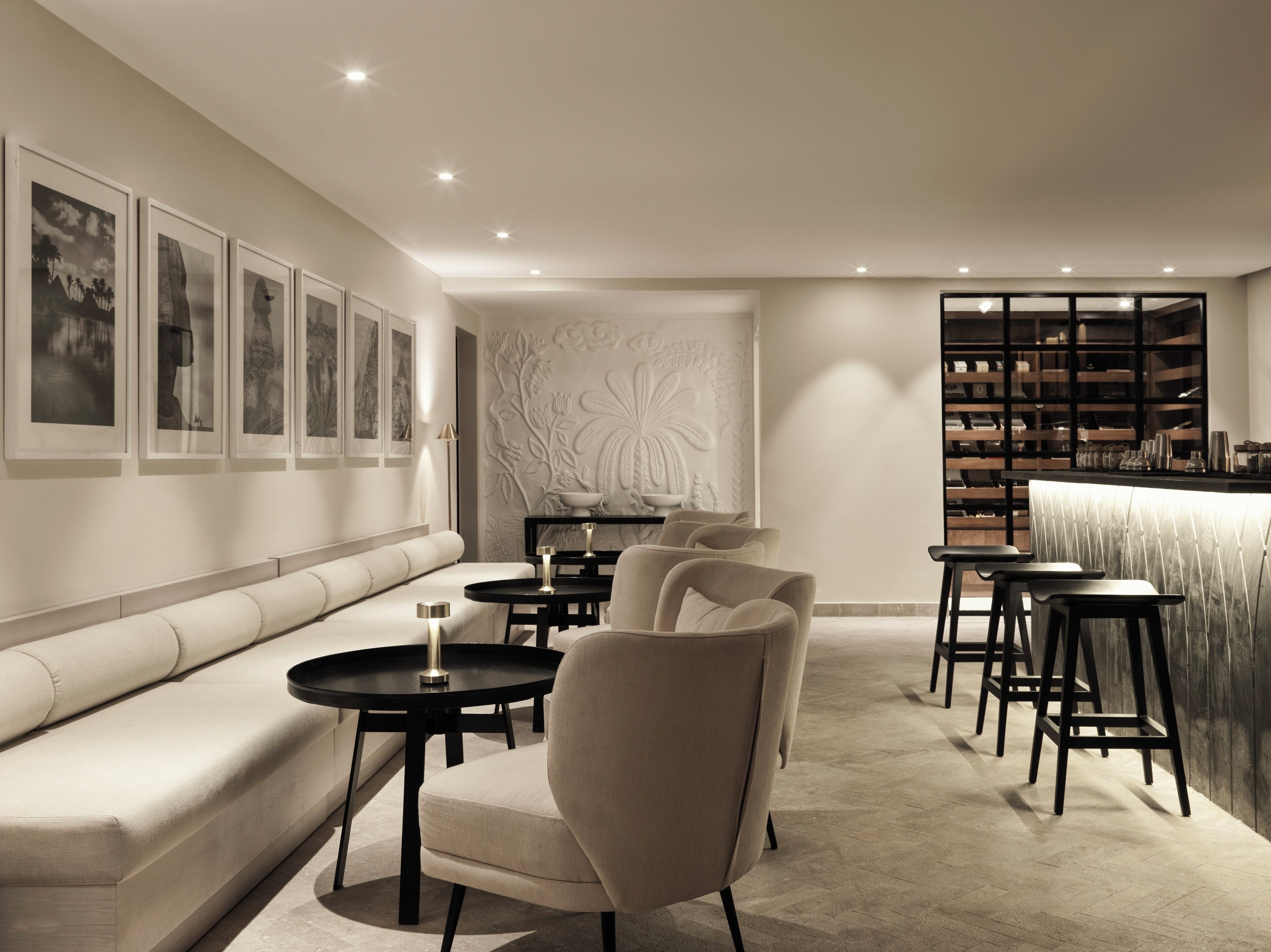A modern lounge area with a sofa, armchairs, and tables, featuring a bar with shelves of bottles and glasses.