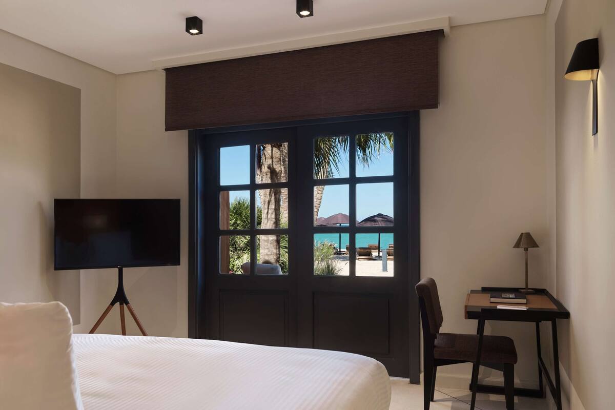 A hotel room with a large bed, a desk with a chair, a TV, and a window with a view of a beach and palm trees.