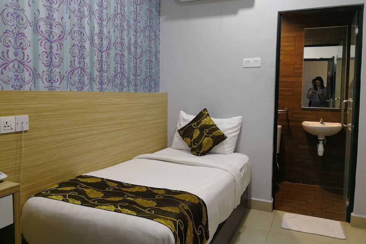 Habitación de hotel con cama, mesita de noche, espejo, baño con lavamanos y unidad de aire acondicionado.
