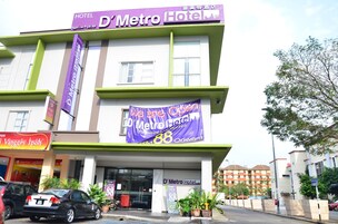 Ein Hotel mit einem violetten Schild für das D'Metro Hotel, einem Banner, das eine Sonderaktion bewirbt, und einem Eingang mit Blumentöpfen.