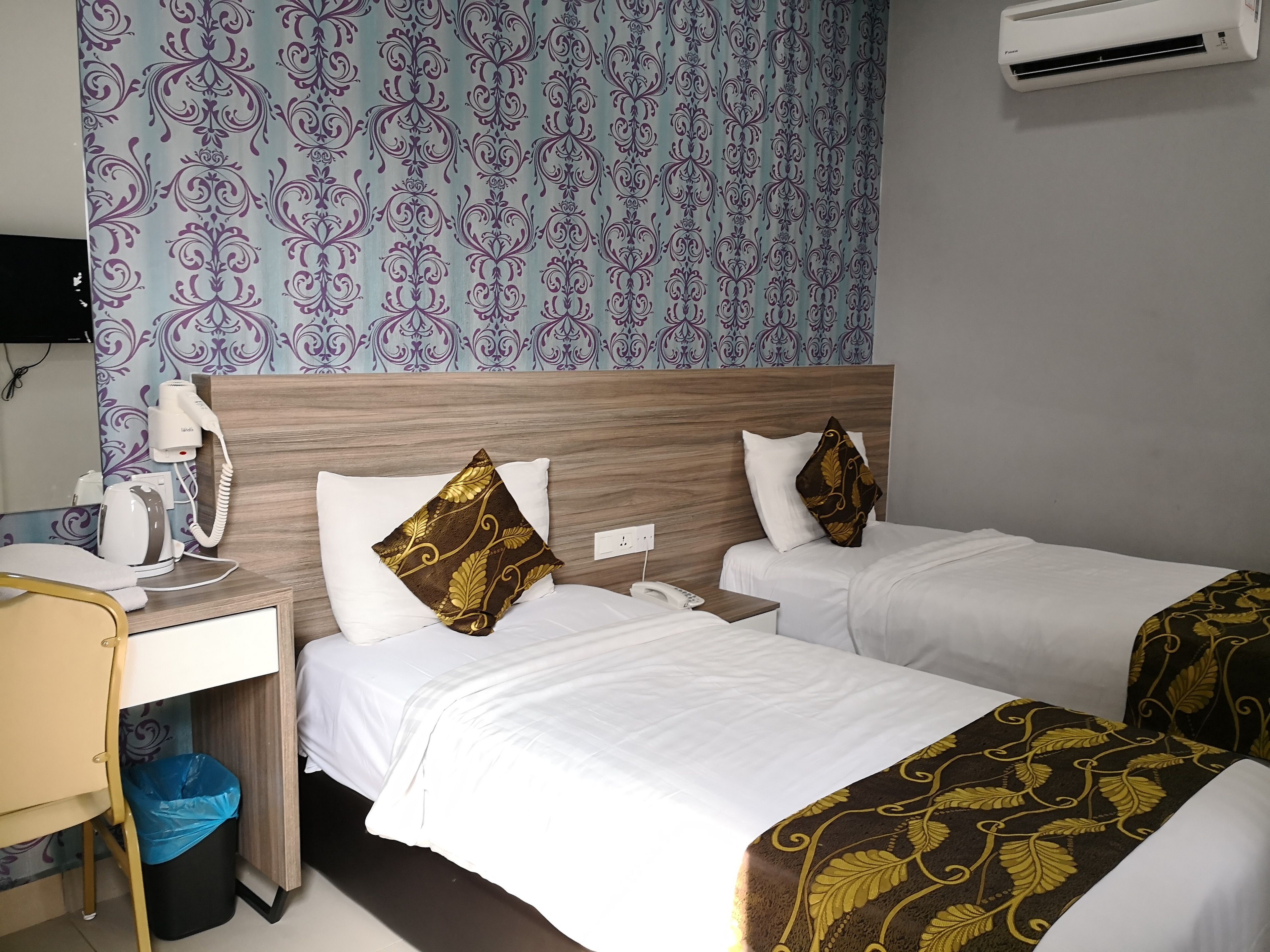A hotel room with two beds, a desk with a chair, a hairdryer, a trash bin, and an air conditioning unit.
