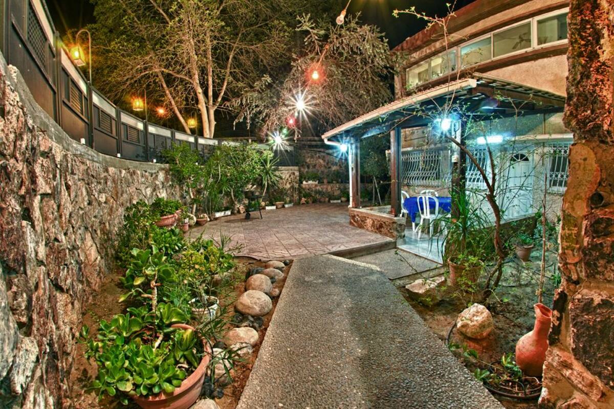 Un patio con paredes de piedra, macetas con plantas, un camino empedrado y una estructura de cristal con sillas azules.