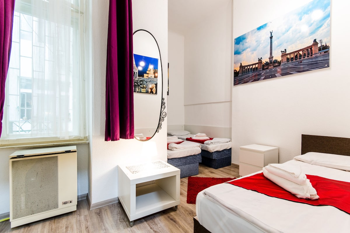 A hotel room with a bed, a desk, a chair, a window with curtains, a heater, a mirror, and a painting on the wall.