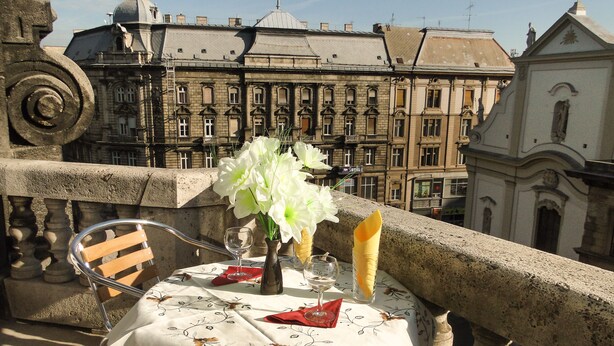 Un balcón con una mesa dispuesta para dos personas, con mantel blanco estampado con motivos florales, un florero con flores blancas y un vaso de agua.