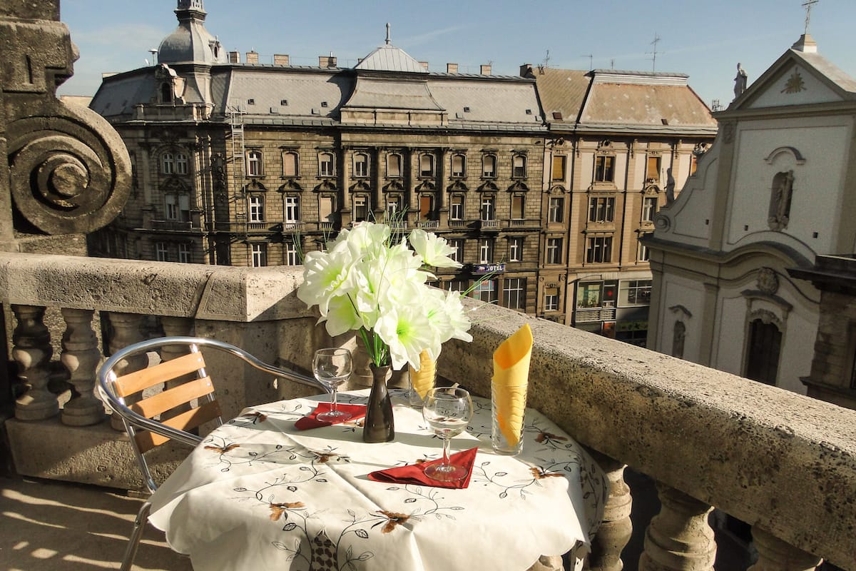 A balcony with a table set for two, featuring a white tablecloth with floral patterns, a vase with white flowers, and a glass of water.