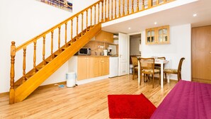 Amplia sala de estar con escalera de madera, cocina con armarios de madera, una mesa de comedor con sillas y un tapete rojo en el suelo.