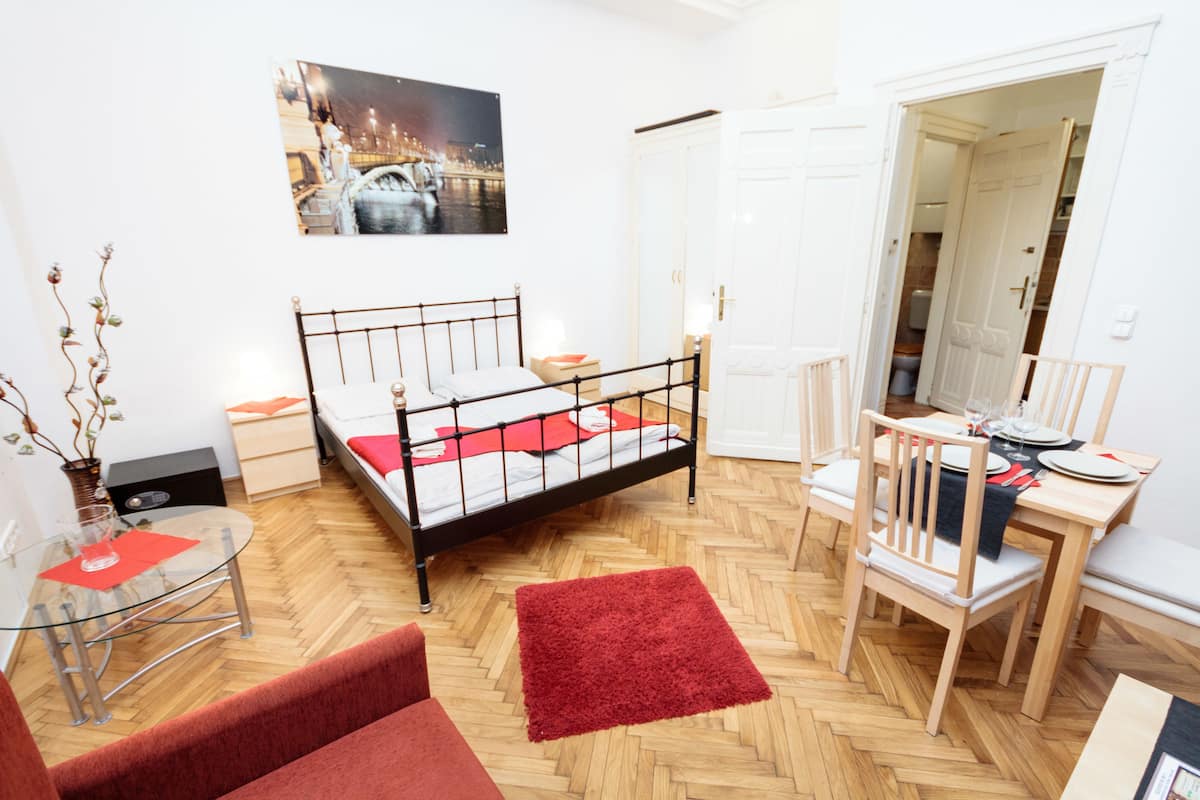 A room with a bed, a dining table with chairs, a small table with a glass top, and a red rug.