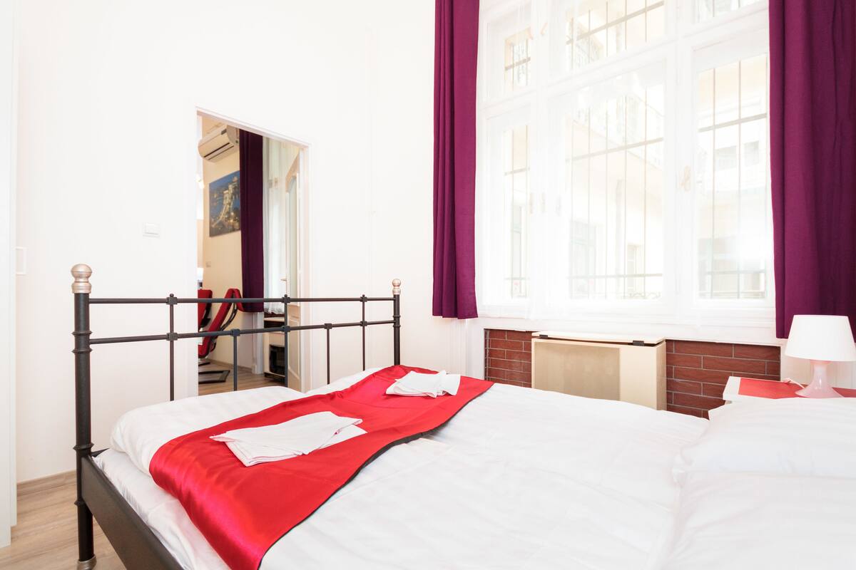 A hotel room with a bed, a window with curtains, a radiator, and a door leading to another room.