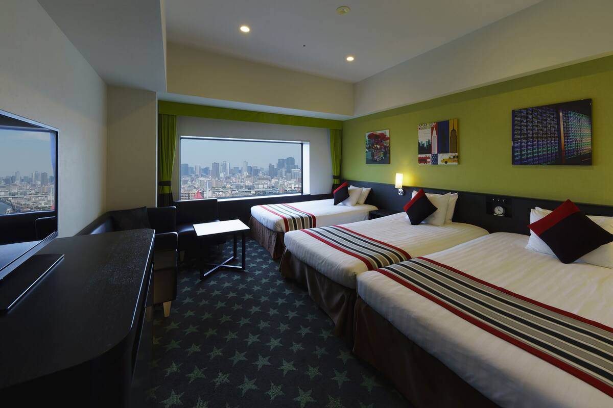 Quarto de hotel com duas camas, uma escrivaninha e uma grande janela com vista para a cidade.