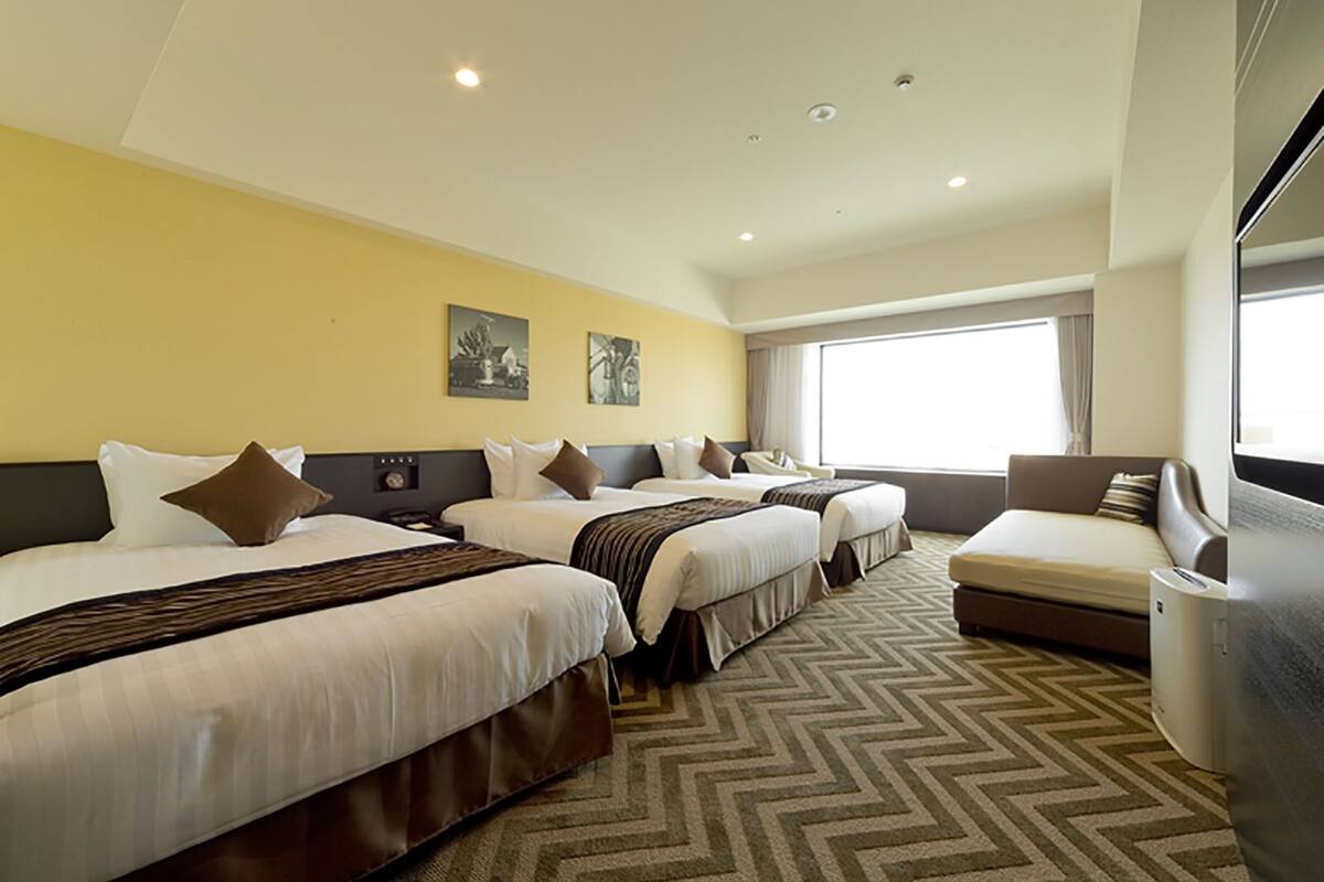 Een hotelkamer met drie bedden, een bank en een groot raam dat natuurlijk licht binnenlaat.
