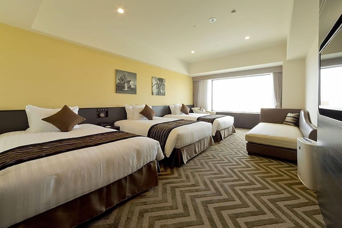 Quarto de hotel com três camas, um sofá, uma janela grande e uma TV fixada na parede.