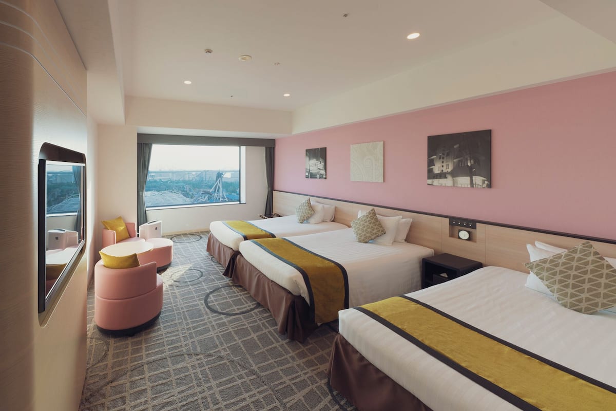 Quarto de hotel com duas camas, uma janela ampla com vista para a cidade, um pequeno espaço de estar e obras de arte nas paredes.