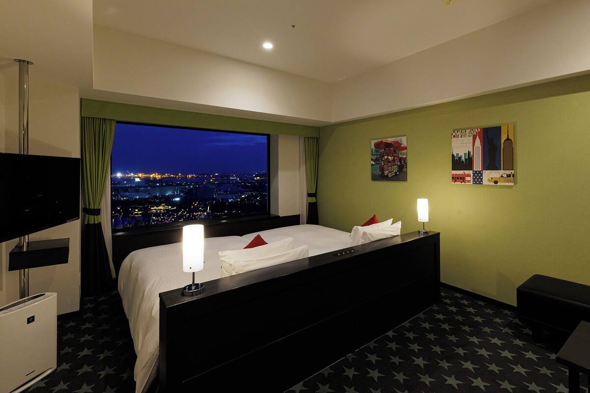 Quarto de hotel com uma grande janela com vista para a cidade à noite, uma cama com lençóis brancos, duas luminárias de cabeceira, uma televisão de tela plana e uma pequena escrivaninha com luminária.