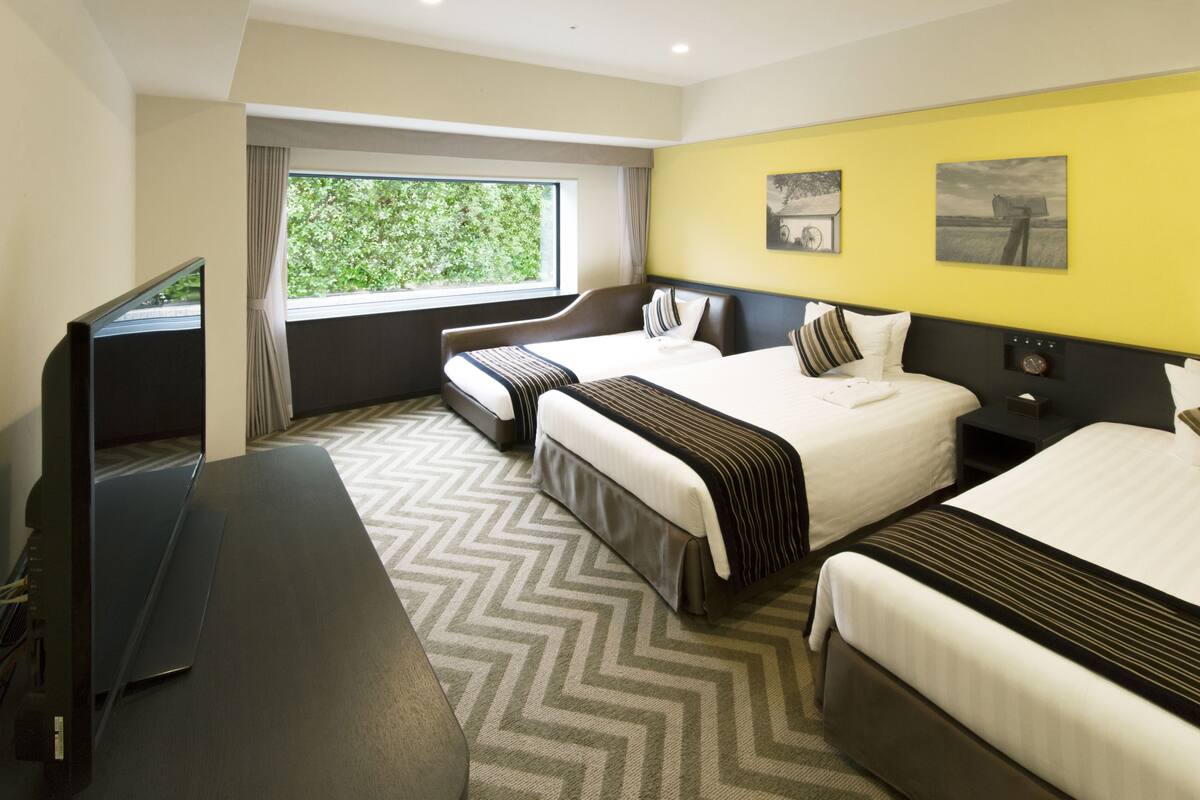 Een hotelkamer met twee bedden, een bureau, een televisie en een groot raam met uitzicht op groen.