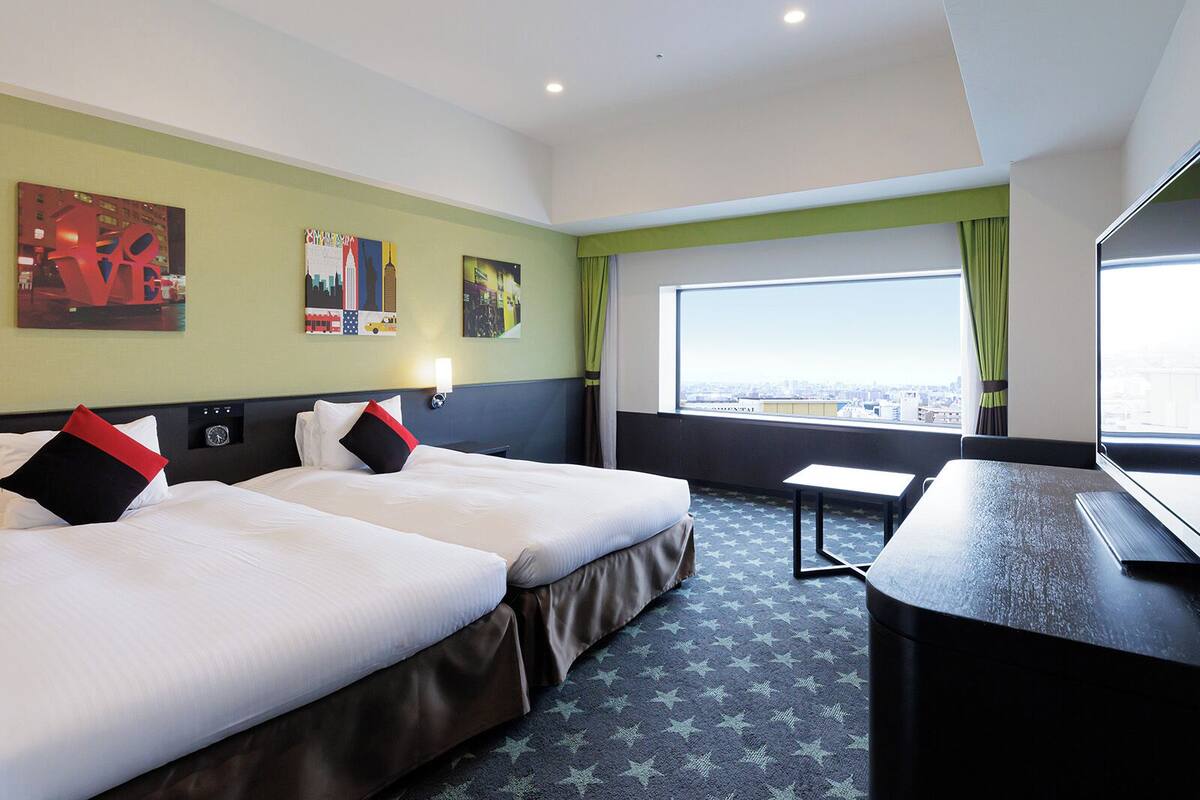 Quarto de hotel com duas camas, uma escrivaninha, televisão e uma janela grande com vista para a cidade.