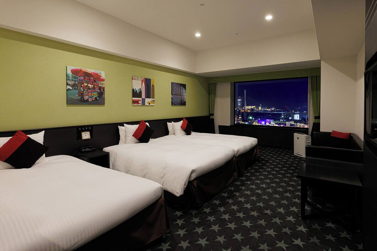 Quarto de hotel com duas camas, uma escrivaninha e uma janela grande com vista para a cidade.
