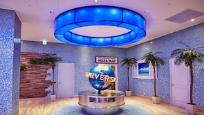 Un hall d’entrée avec un mur décoré d’une mosaïque bleue, un mur recouvert de panneaux de bois, une enseigne indiquant « Wolfgang Puck Pizza Bar » et un globe terrestre sur lequel est inscrit le mot « UNIVERSAL ».
