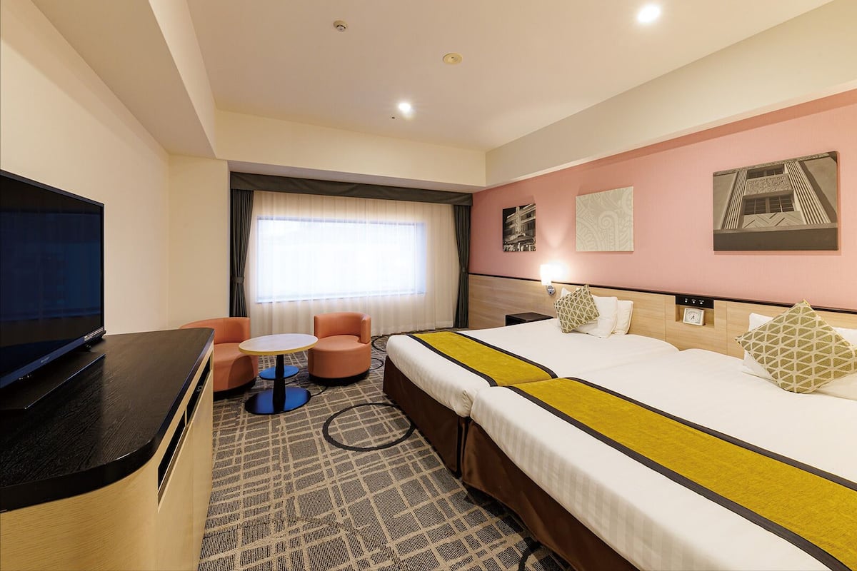 Een hotelkamer met een bed, een televisie, een klein tafeltje, twee stoelen, een raam met gordijnen en kunst aan de muren.