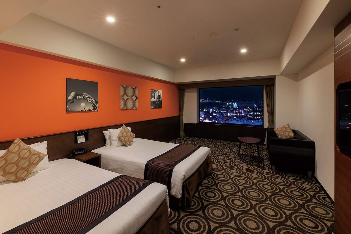 Een hotelkamer met twee bedden, een bureau en een groot raam met uitzicht op de stad.