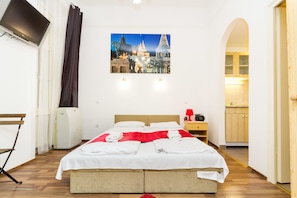 A hotel room with a bed, a flat-screen TV, a painting of a cityscape, and a view of the kitchen.