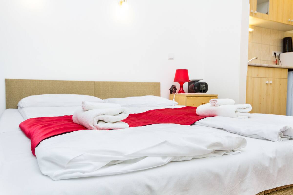 A neatly made bed with white and red bedding, a wooden headboard, and a small bedside table with a red lamp and a black object.