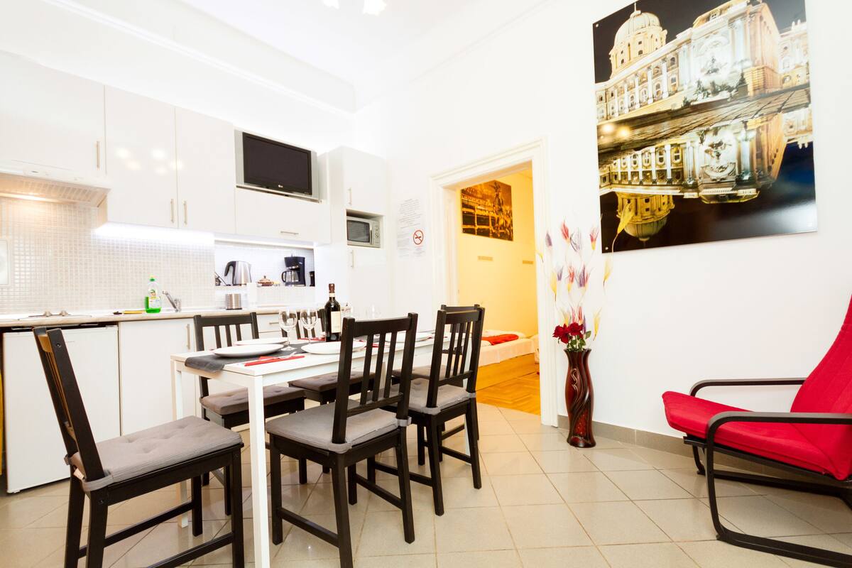 A modern kitchen with white cabinets, a dining table with chairs, a red chair, and a wall-mounted TV.