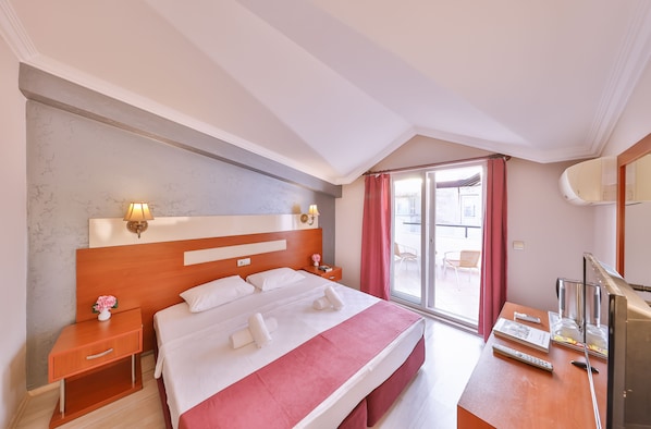 A hotel room with a large bed, a nightstand with a lamp, a desk with a TV, and a sliding glass door to the outside.
