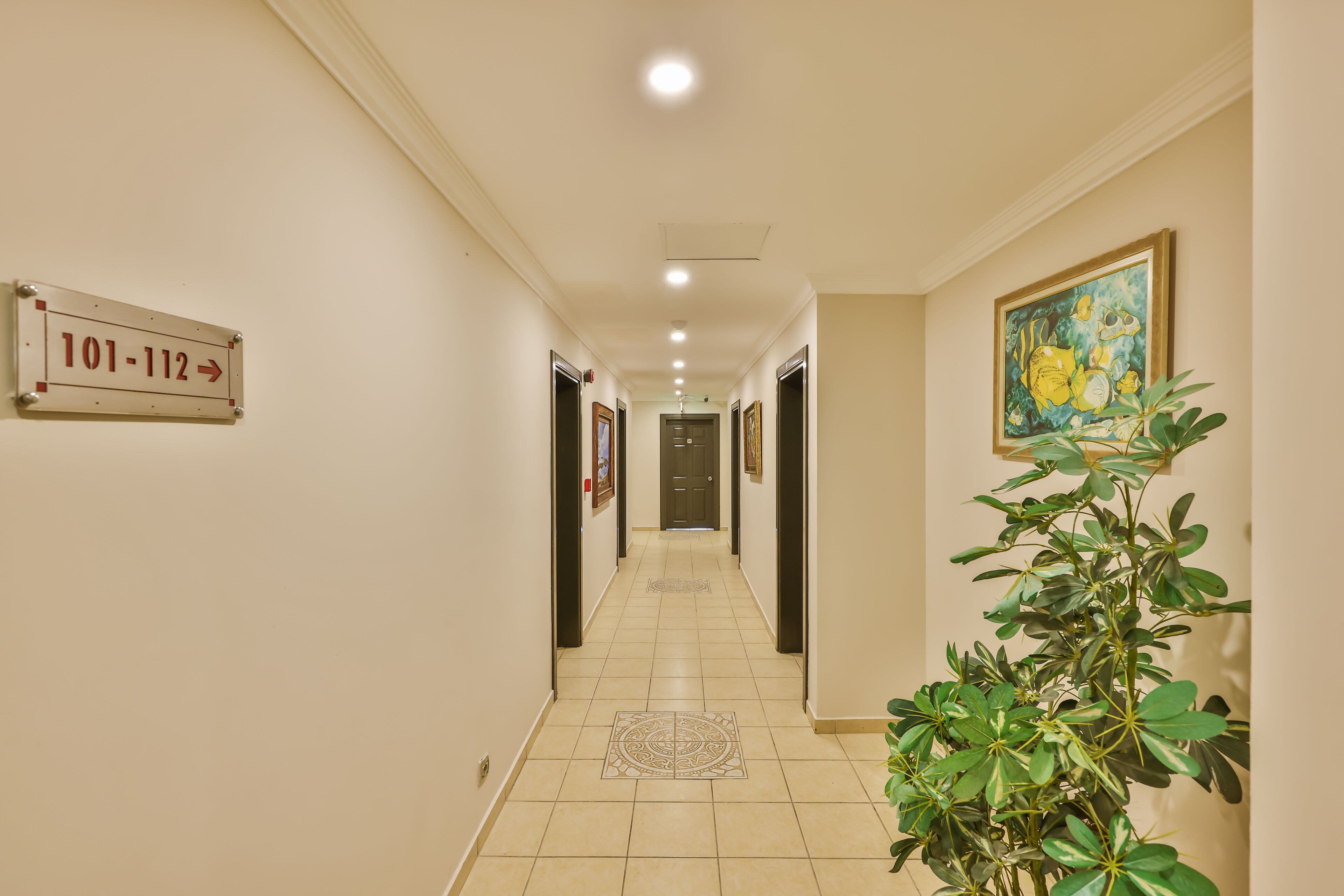 Un couloir d’hôtel avec un panneau indiquant les numéros de chambre 101 à 112, un sol carrelé et une plante sur la droite.
