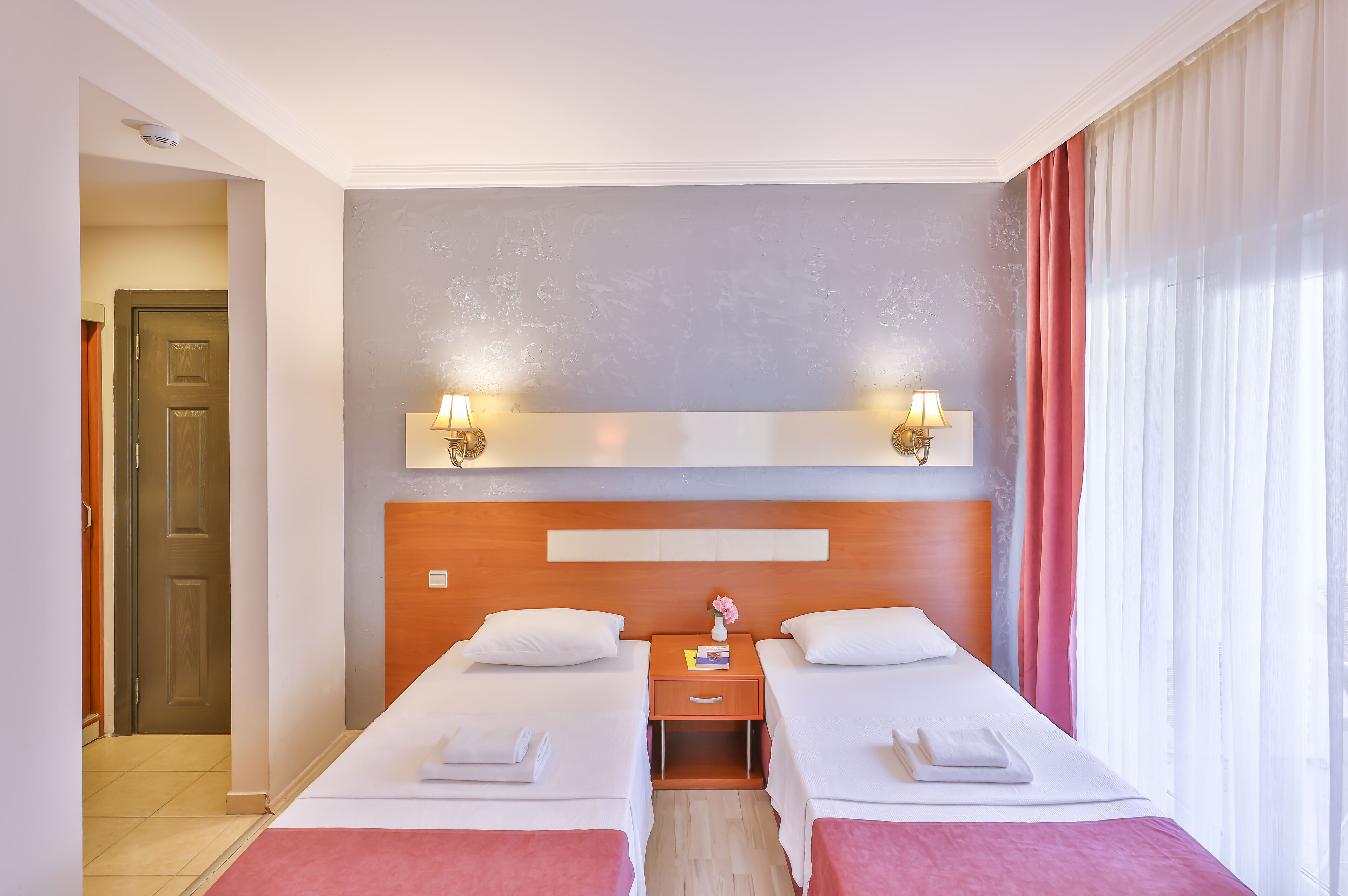 Habitación de hotel con dos camas, cabecero de madera, mesita de noche con lámpara y vista al exterior a través de una ventana con cortinas.