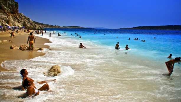 Una playa con agua azul cristalina, gente nadando y tomando el sol, y acantilados rocosos al fondo.
