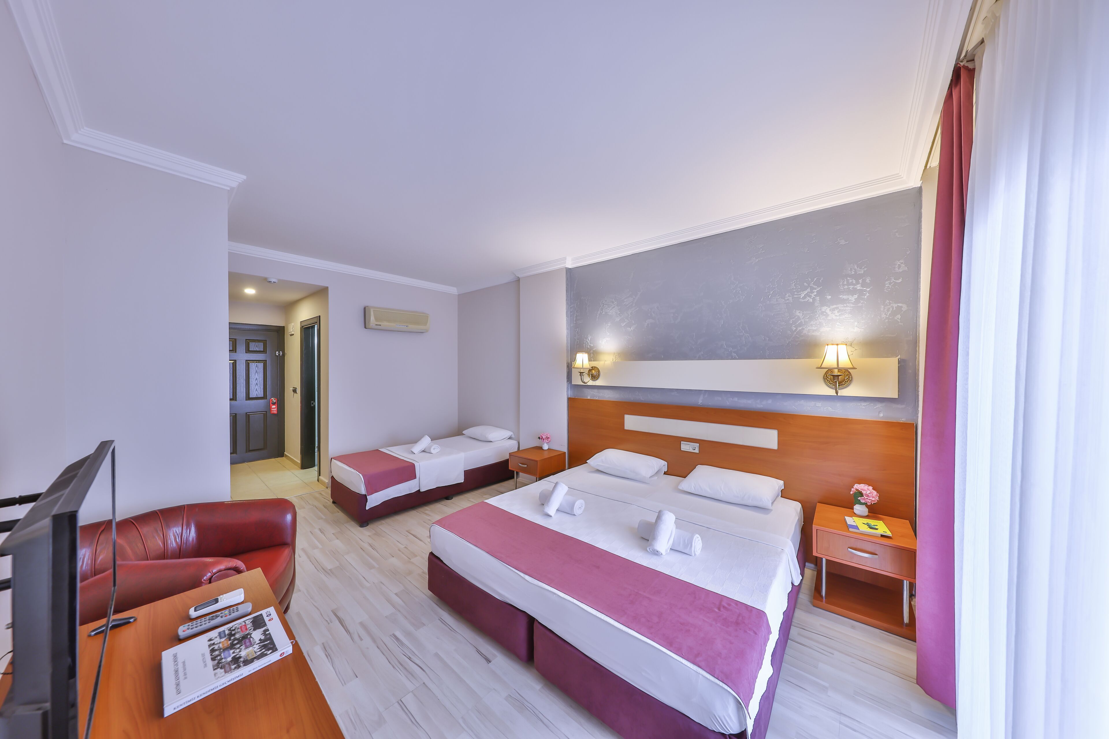Habitación de hotel con una cama grande, una cama más pequeña, un televisor, un escritorio y un sofá de cuero rojo.