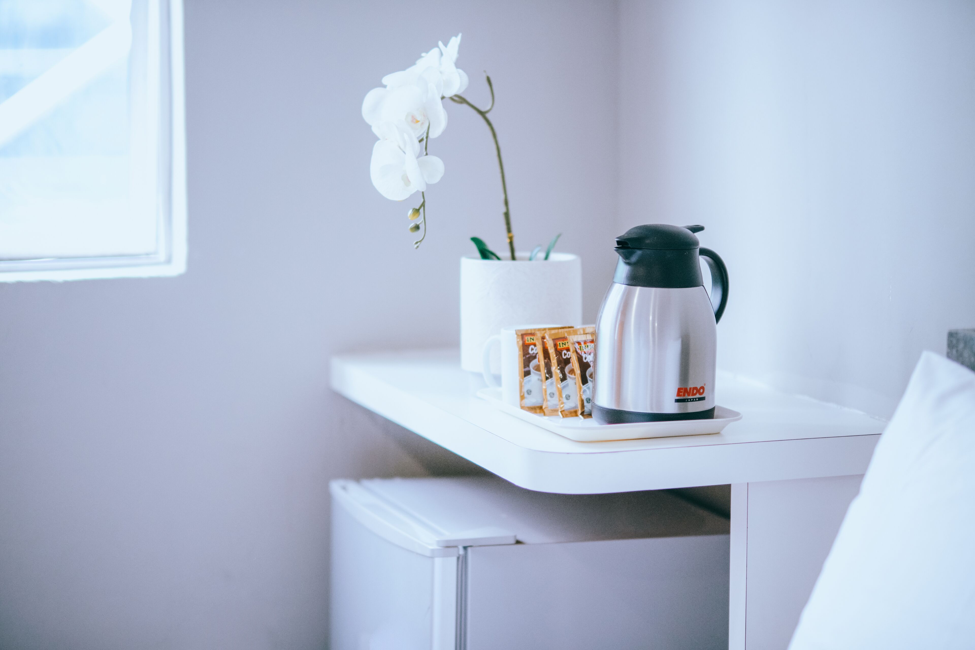 Uma mesa branca com uma orquídea branca, uma chávena branca e uma cafeteira de prata.