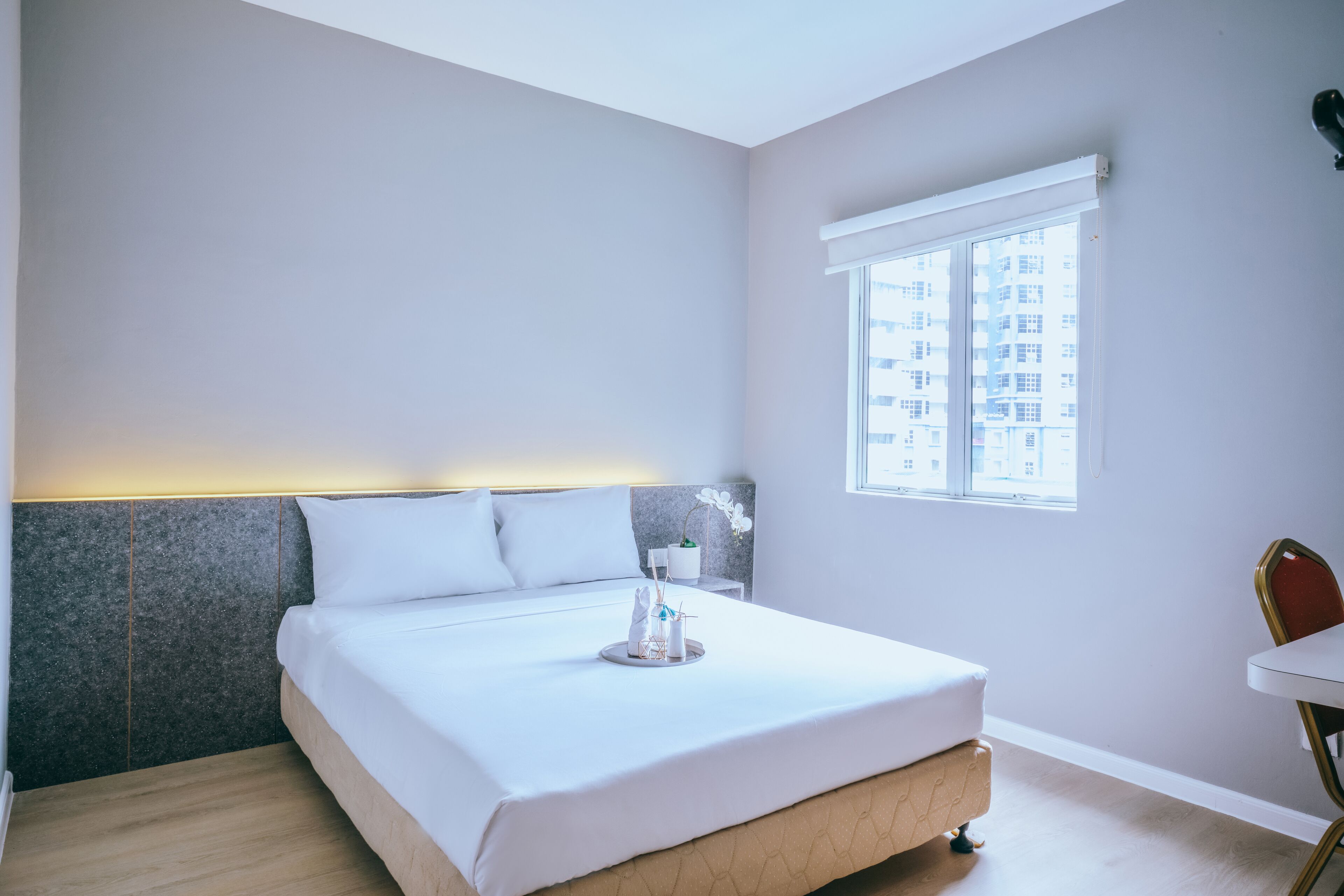 A modern hotel room with a large bed, a small bedside table, a window with a view of buildings, and a chair.