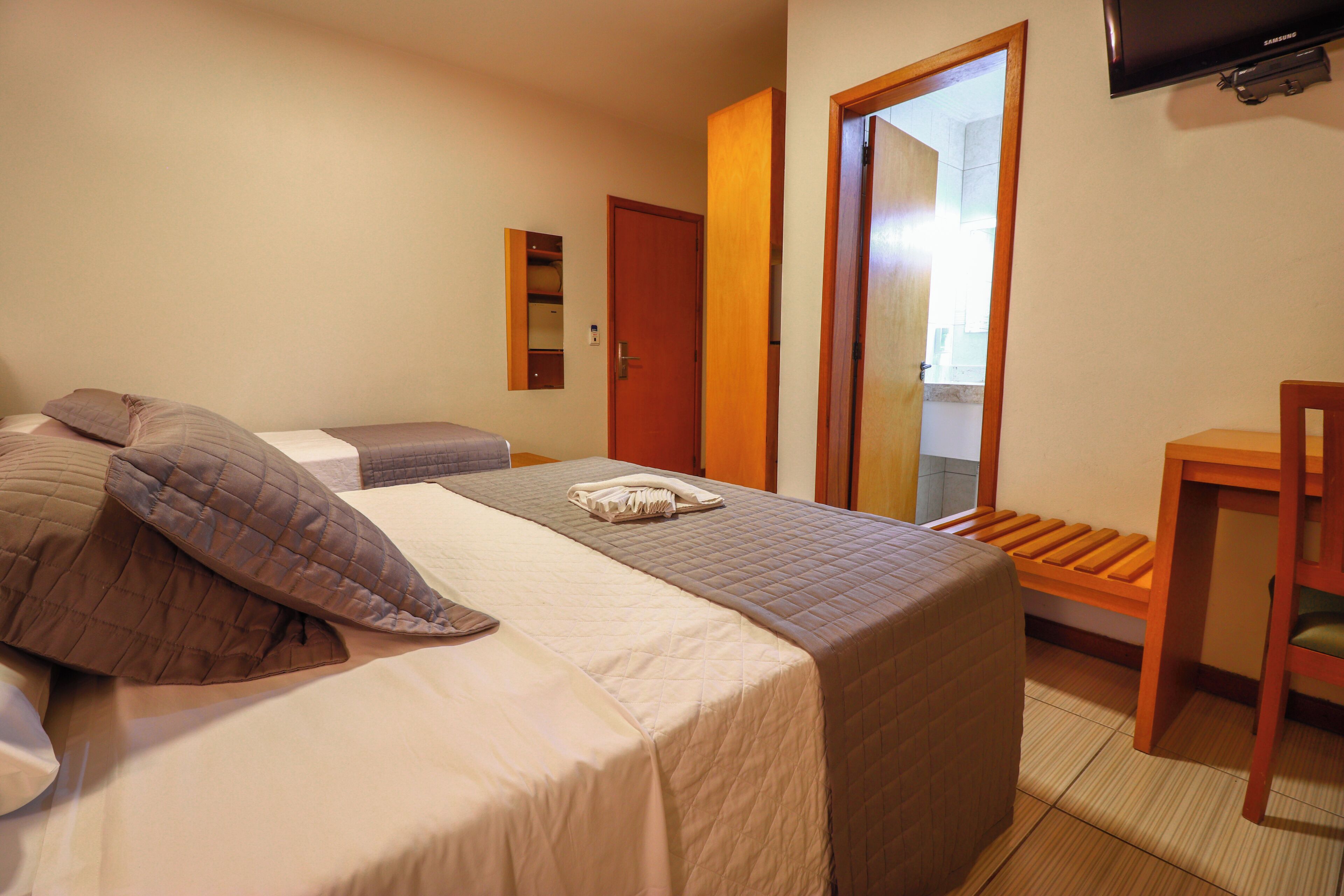 A hotel room with a bed, a wooden bench, a television, and a door leading to a bathroom.