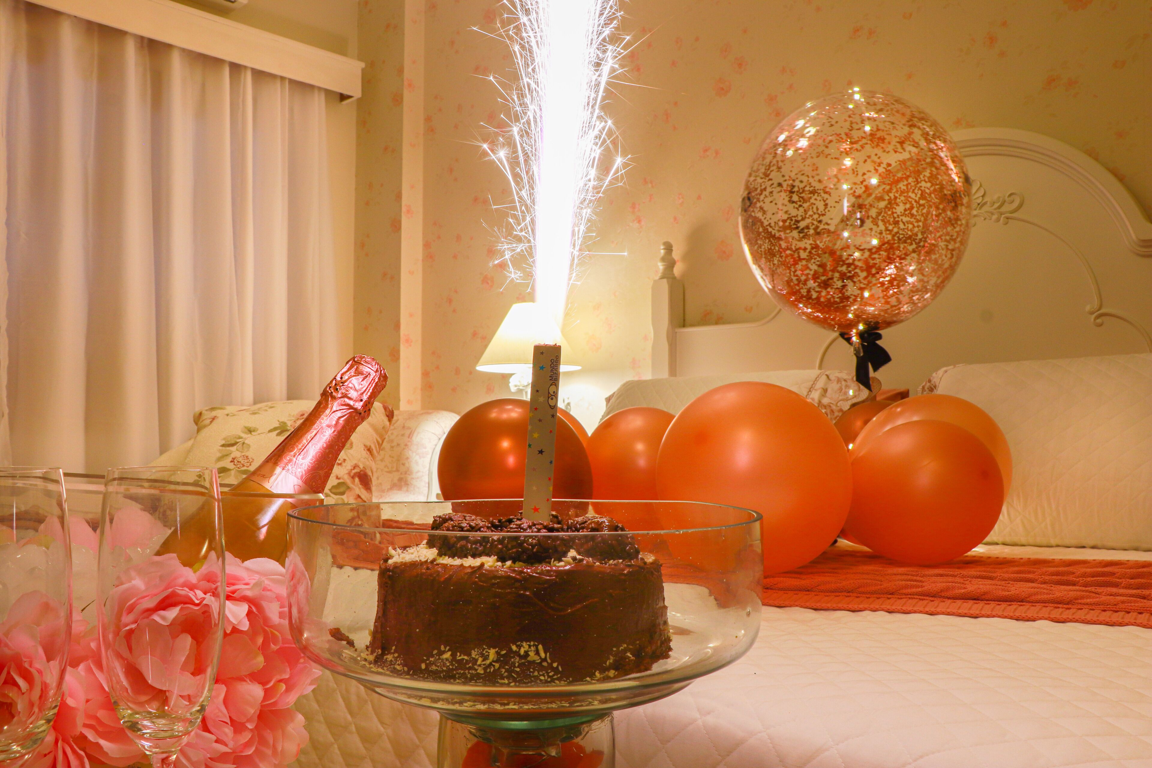 Una habitación con una cama, un pastelero de cristal con un pastel de chocolate, globos y una botella de champán.