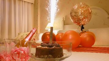 Una habitación con una cama, un pastelero de cristal con un pastel de chocolate, globos y una botella de champán.