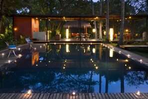 A serene outdoor pool area with a wooden deck, lounge chairs, and a covered seating area with a thatched roof.