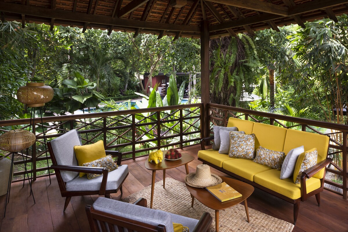 A covered outdoor seating area with a yellow sofa, wooden chairs, and a woven table.