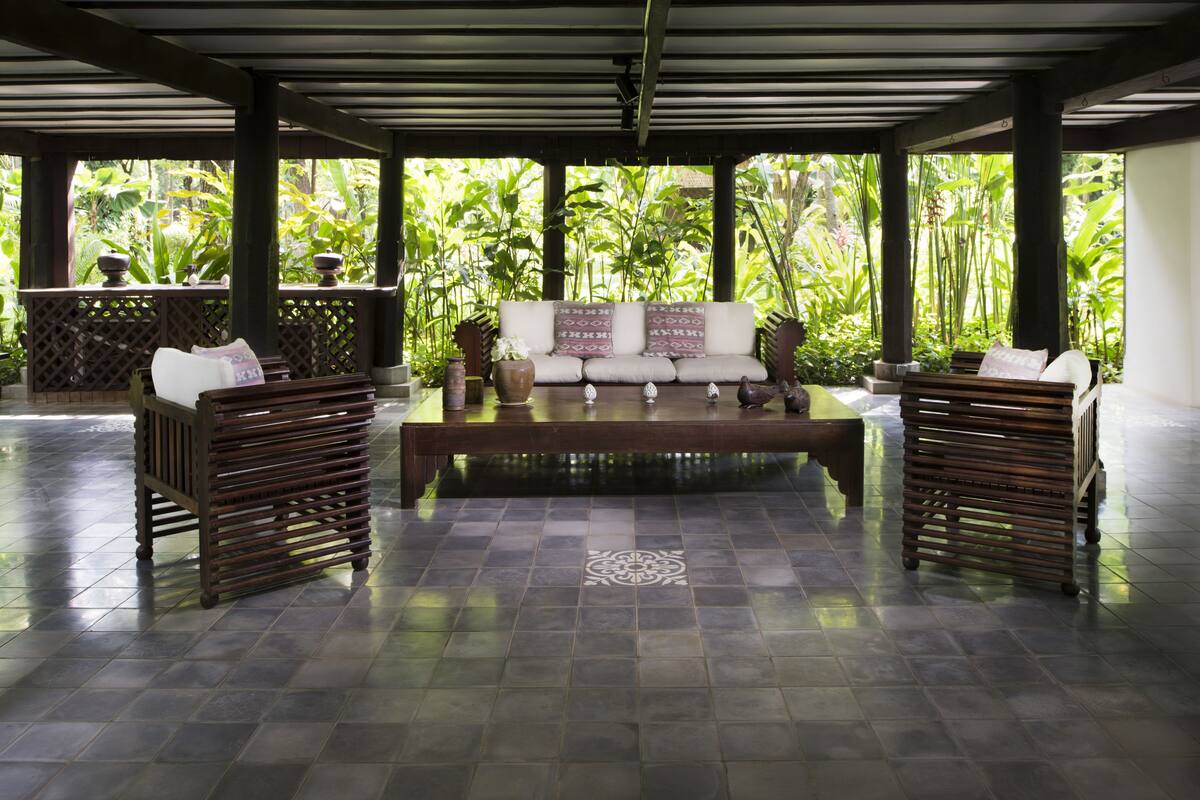 A covered outdoor seating area with a sofa and two wooden benches, surrounded by lush greenery.