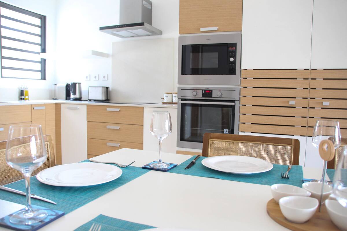 Una cocina moderna con electrodomésticos integrados, una mesa de comedor para dos personas con un mantel individual y una ventana con persianas.