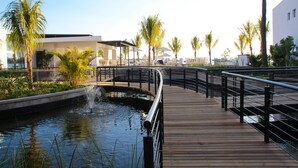 Un paseo marítimo de madera sobre una fuente de agua, con palmeras y edificios modernos al fondo.